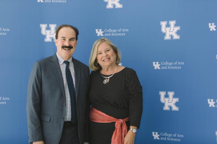 A man and woman in formal attire stand together in front of a backdrop featuring the University of Kentucky College of Arts and Sciences logo.