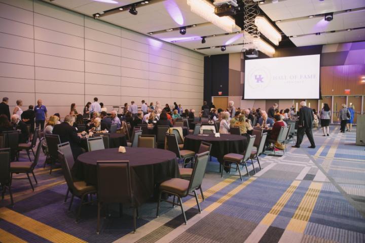 A large room with several round tables and a crowd of people, with a projector screen visible on the wall.