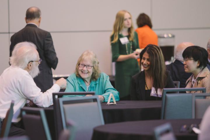 A group of people sit at a table in a conference room chatting and smiling.