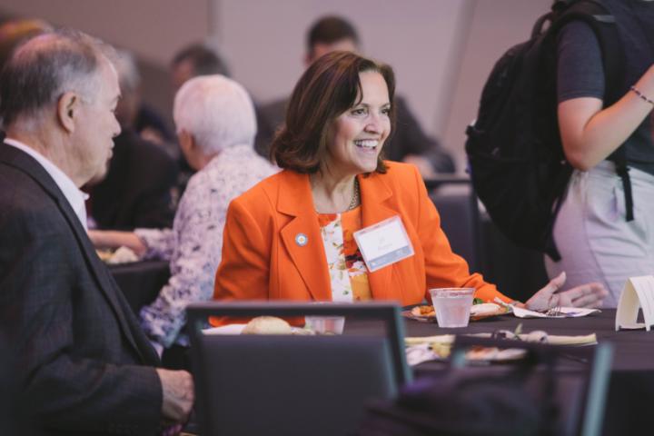 A woman in an orange blazer smiles as she interacts with a few people at a table.