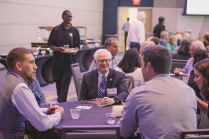 A large group of people sit at several tables in a large room, talking and socializing while a few staff members serve food in the background.
