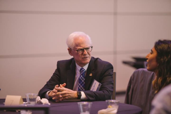 A man in a suit sits at a table with several other people, engaged in conversation.