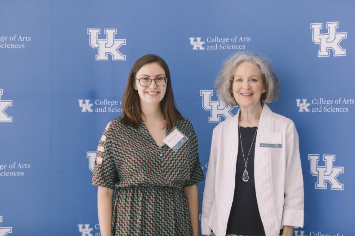 Two women stand together in front of a blue backdrop featuring the University of Kentucky College of Arts and Sciences logo.