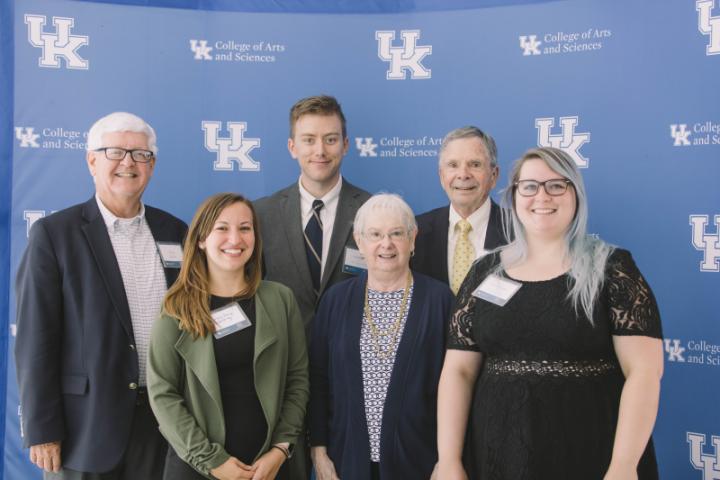 A group of six people stand in front of a blue backdrop with logos from the University of Kentucky College of Arts and Sciences.