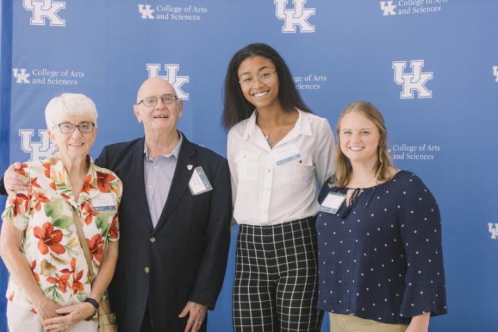 Four people stand together, posing in front of a backdrop featuring the University of Kentucky College of Arts and Sciences logo.