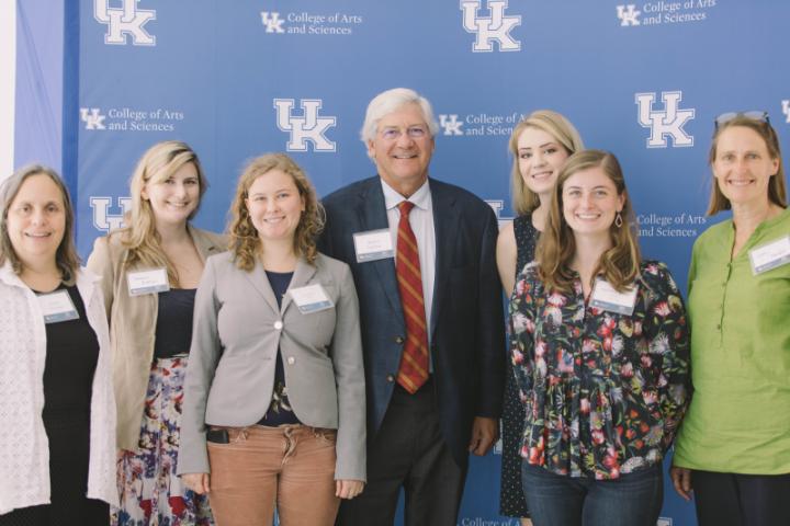 A group of people, including a man in a suit and tie, stand together in front of a blue backdrop with the University of Kentucky logo.