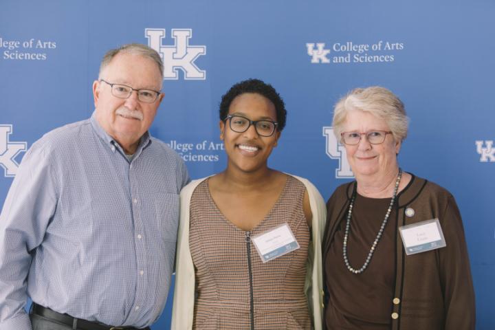 A man and two women stand together, smiling and posing in front of a backdrop bearing the University of Kentucky logo.