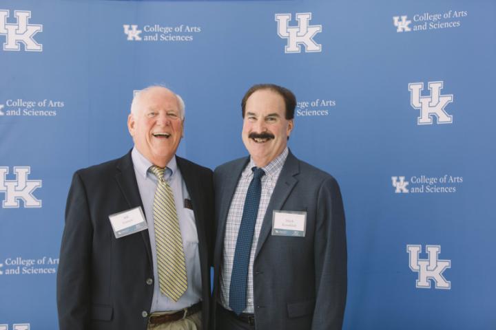 Two men in business attire stand in front of a blue backdrop with the University of Kentucky logo and College of Arts and Sciences text.