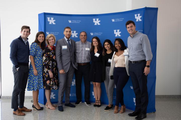 A group of people in business casual attire stand in front of a blue backdrop with University of Kentucky logos.