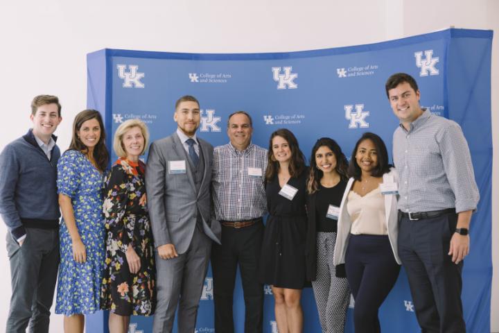 A group of people stand together, smiling, in front of a backdrop bearing the University of Kentucky logo and College of Arts and Sciences name.
