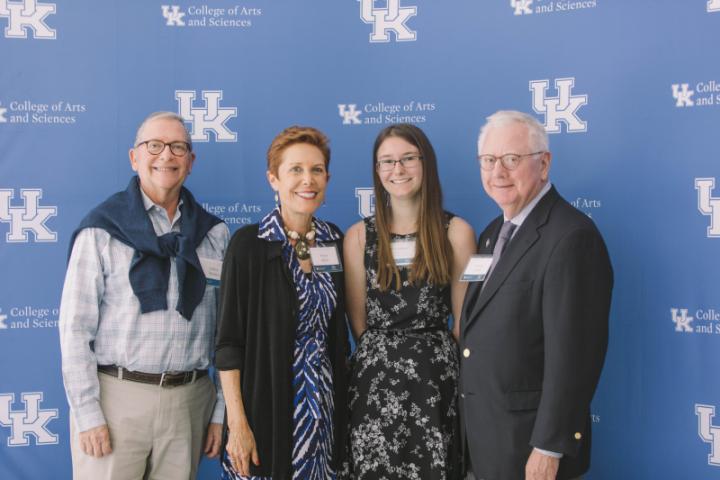 A group of four people stand together, smiling, in front of a backdrop with the University of Kentucky College of Arts and Sciences logo.