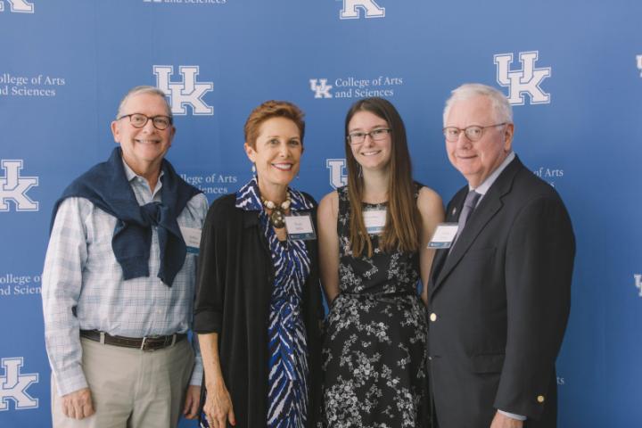 Four people stand together, posing for a photo in front of a backdrop with the University of Kentucky logo.
