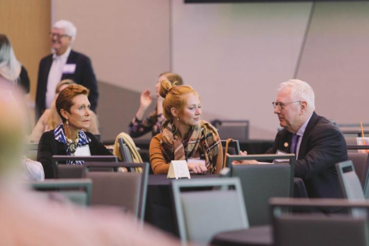 Several people are seated at desks in a large room, engaged in conversation.