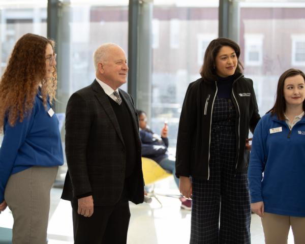 A group of people, including a few wearing blue sweaters with name tags, stand together in a large room with a view of a brick building outside.