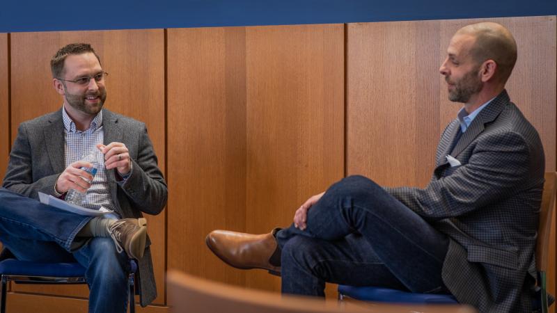 A man converses with another man while seated in chairs.