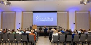 Wide view of speaker on stage with "Circle of Giving" screen.