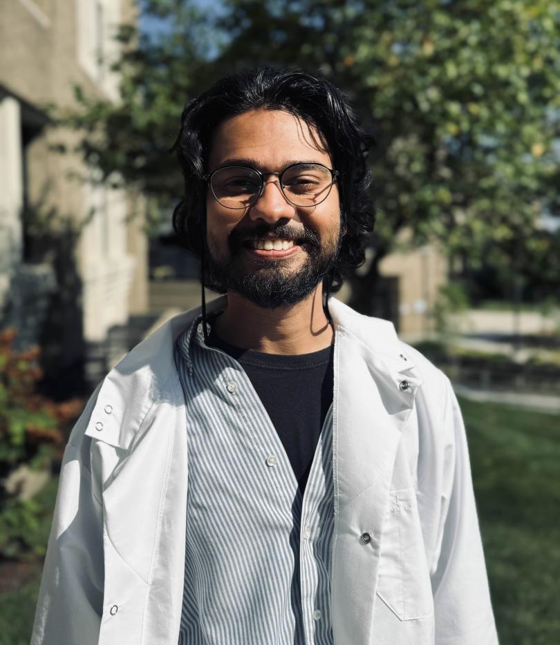 Photo of Dr. D'Souza smiling in a white lab coat. In the background you can see blurred green leaves from a tree in the back right corner and the side of a building in the back left corner.
