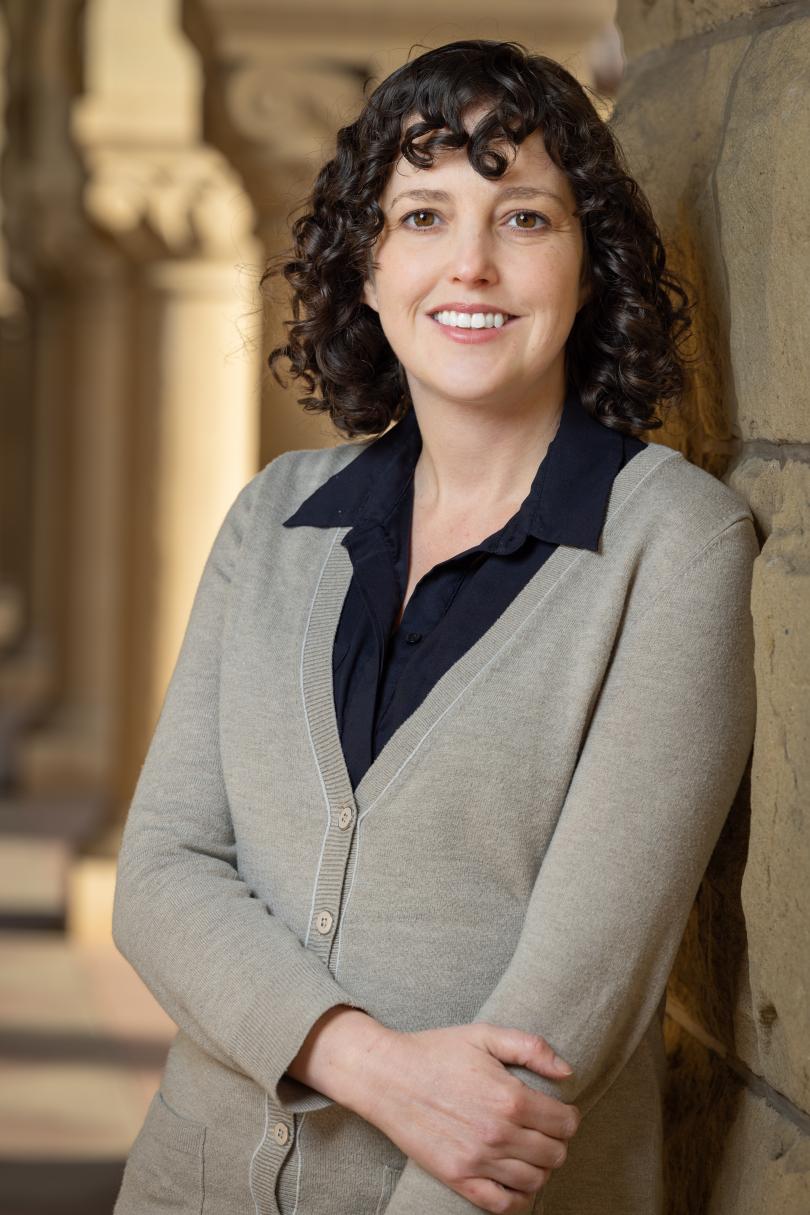 Dr. Molly Schumer leaned up against a stone wall with one arm across her waist holding the other arm while smiling.